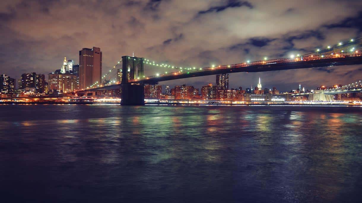 Night view of the Brooklyn Bridge with Manhattan skyline lights reflecting on the East River