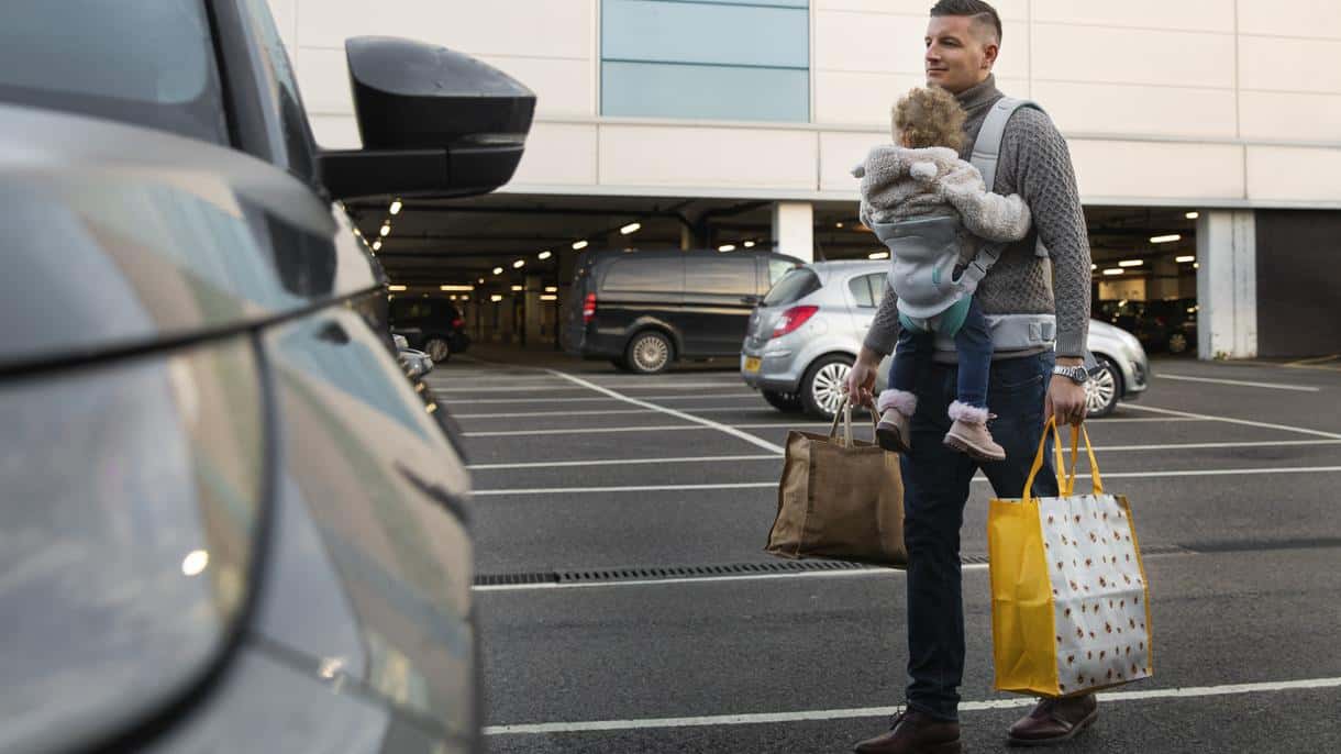 A father with a child in a sling and bags goes to a rented car in the airport parking lot.