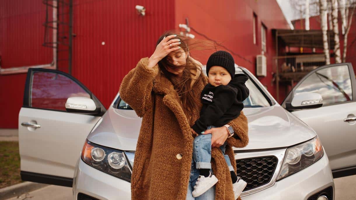 A mom holds a baby in her arms next to a silver sedan, a comfortable car for a mom and child in the city.